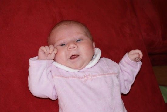 Ein Babymädchen liegt auf einer roten Decke, trägt ein rosa Outfit und lächelt mit erhobenen Händen.
