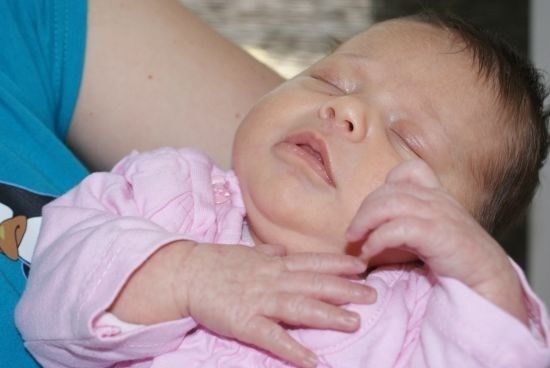 Ein Babymädchen in einem rosa Outfit schläft, während es von jemandem in einem blauen Shirt gehalten wird.