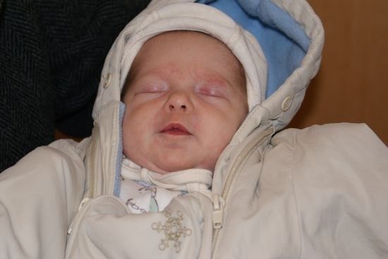Ein neugeborenes Baby schläft, gekleidet in einem weißen und blauen Mantel mit Kapuze. Das Baby trägt ein weißes Lätzchen mit Schneeflockendesign.