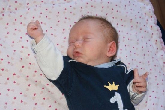 Ein Babyjunge schläft friedlich auf einer Decke mit Herzmuster und trägt ein blaues Shirt mit gelber Krone und der Nummer 1.