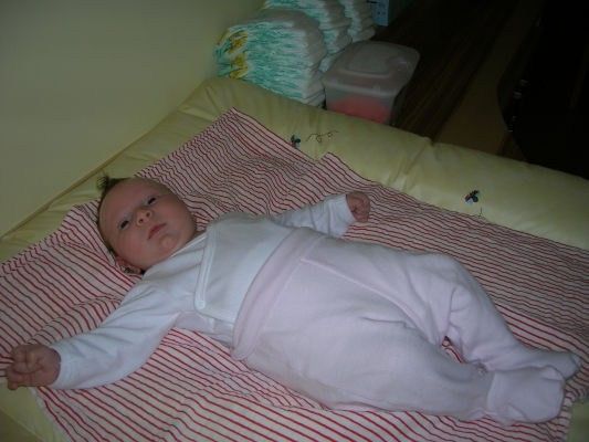 Ein Baby in weißer Kleidung liegt auf einer rot-weiß gestreiften Decke auf einem Bett. Hinter dem Baby befinden sich mehrere Stapel von Windeln.