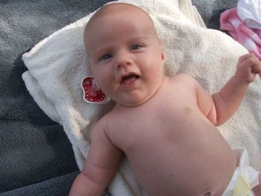 Ein Baby liegt auf einem weißen Handtuch, trägt einen Windel und einen Schnuller und lächelt in die Kamera.