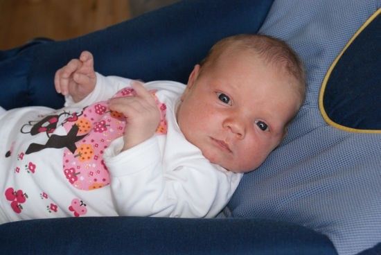 Ein Babymädchen liegt auf einem blauen Kissen und trägt ein weißes Shirt mit rosa Blumendesigns. Sie schaut neugierig direkt in die Kamera.