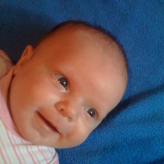 Ein Nahaufnahme eines lächelnden Babys mit blauen Augen, das auf einer blauen Decke liegt. Das Baby trägt ein rosa-weiß gestreiftes Shirt.