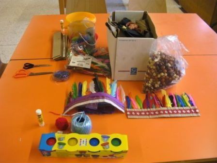Ein orangefarbener Tisch zeigt eine Vielzahl von Bastelmaterialien, darunter eine Schachtel mit Perlen, bunte Federn, eine gelbe Schachtel mit einem Puzzle, Scheren, einen gelben Eimer und andere verschiedene Gegenstände.