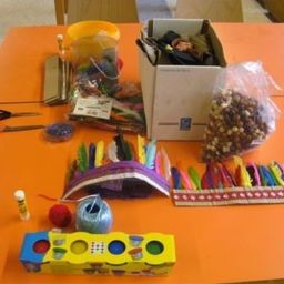 Ein orangefarbener Tisch zeigt eine Vielzahl von Bastelmaterialien, darunter eine Schachtel mit Perlen, bunte Federn, eine gelbe Schachtel mit einem Puzzle, Scheren, einen gelben Eimer und andere verschiedene Gegenstände.