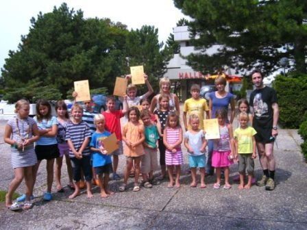 Eine Gruppe von Kindern und Erwachsenen, die gelbe Papiere halten, posiert für ein Foto vor einem Gebäude mit Bäumen im Hintergrund.