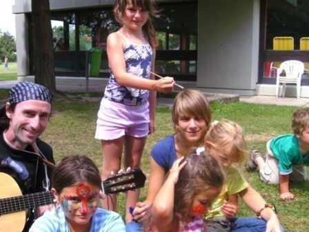 Eine Gruppe von Kindern und einem Erwachsenen sitzen auf dem Gras. Der Erwachsene spielt Gitarre und die Kinder haben Gesichtsbemalung. Sie lächeln und posieren für ein Foto.