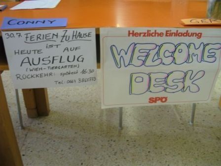 Ein Schreibtisch mit zwei Schildern, eines auf Deutsch mit 'Herzliche Einladung' und 'WELCOME DESK', das andere auf Englisch mit einer Telefonnummer und 'ROCKKEHR'. Der Schreibtisch steht auf einem gefliesten Boden.