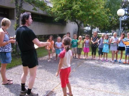 Ein Mann in schwarzer Kleidung führt eine Gruppe von Kindern in einem Kreis auf einem gepflasterten Bereich an, wobei Erwachsene in der Nähe stehen und ein Gebäude im Hintergrund zu sehen ist.