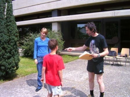 Ein Mann in einem schwarzen T-Shirt und schwarzen Shorts hält ein Stück Papier und zeigt auf einen Jungen in einem roten T-Shirt und weißen Shorts, während eine Frau in einem blauen Langarmshirt steht.