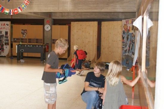 Drei Kinder in einem Raum mit Holzwänden und einer Uhr an der Wand. Ein Junge hält eine Tasche, und zwei andere schauen auf eine Glaswand.