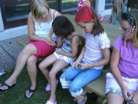 Fünf junge Mädchen sitzen auf einer Holzbank und beschäftigen sich mit einer Bastelaktivität. Eines der Mädchen trägt eine rote Bandana, ein anderes ein rosa Kleid und alle haben Kopfhörer auf.