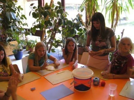 Eine Gruppe junger Mädchen sitzt um einen Tisch herum und ist mit einer Frau vor ihnen, die sie anleitet, in einer Bastelaktivität beschäftigt. Der Tisch ist mit orangefarbenem Stoff bedeckt und hat verschiedene Bastelmaterialien.