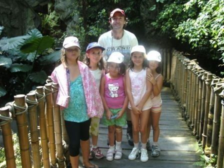 Ein Mann und fünf Kinder stehen auf einer Holzbrücke im Wald, alle tragen Hüte und lächeln für ein Foto.