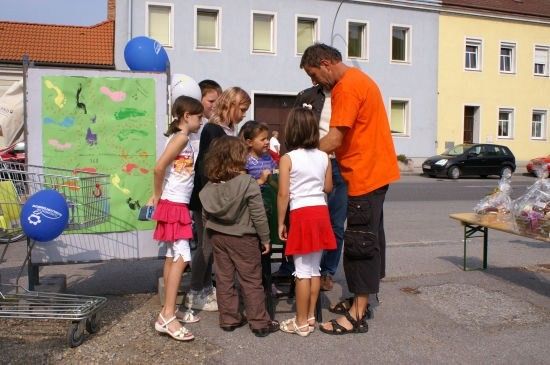 Eine Gruppe von Kindern und Erwachsenen steht vor einem Gebäude mit einer bunten Malerei und Luftballons. Ein Mann in einem orangefarbenen T-Shirt spricht mit den Kindern.
