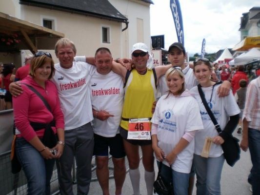 Eine Gruppe von Menschen, wahrscheinlich Marathonteilnehmer, stehen lächelnd für ein Foto zusammen. Sie tragen weiße T-Shirts mit Nummern und ein Mann hat ein gelbes T-Shirt. Hinter ihnen sind Zelte und Gebäude.