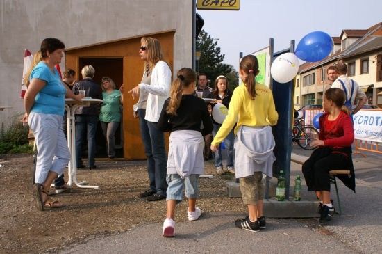 Eine Gruppe von Menschen, darunter Frauen und Kinder, steht vor einem Gebäude mit einem Banner und Luftballons. Einige tragen Turnschuhe und haben Taschen dabei.