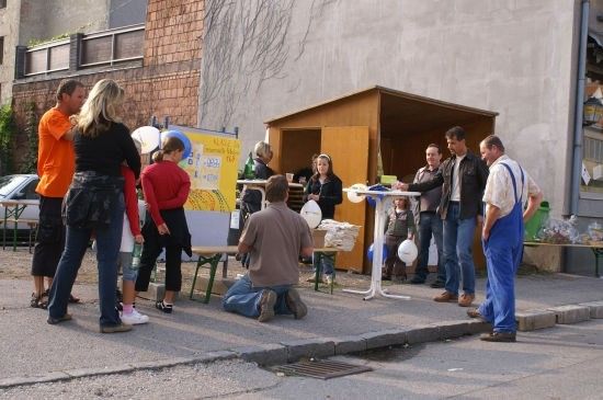 Eine Gruppe von Menschen versammelt sich um eine kleine Holzbude auf einem Bürgersteig, einige knien und andere stehen. Es gibt Luftballons und einen Tisch mit einem gelben Schild.