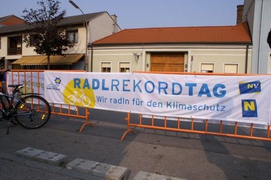 Ein Banner zur Förderung des 'Radrekordtags' ist über orange Barrieren auf einer Straße gespannt, mit einem Fahrrad in der Nähe. Das Banner zeigt ein Fahrrad-Symbol und Text in Blau und Gelb.