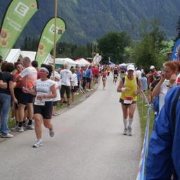 Zwei Läufer nehmen an einem Marathon teil, mit Zuschauern und Bannern am Straßenrand, Berge im Hintergrund.