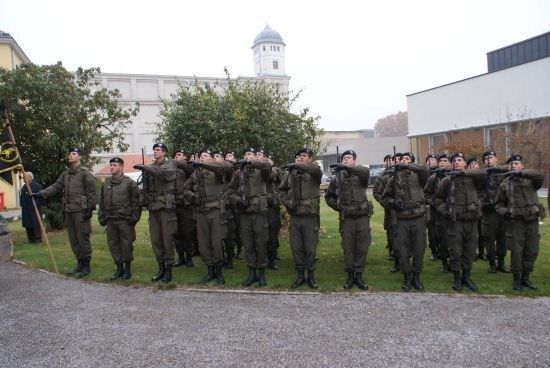 Eine Gruppe von Soldaten in Uniform, mit Gewehren, steht in Formation auf einem Rasen vor einem Gebäude.