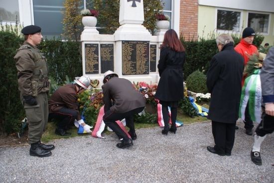 Eine Gruppe von Menschen, einschließlich Militärangehöriger, versammelt sich um ein Denkmal und legt Blumen und Kränze auf den Boden.