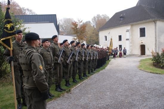 Eine Reihe von Militärangehörigen in Uniform steht in Formation vor einem Gebäude mit einer Fahne auf der linken Seite.