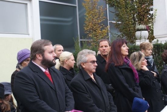 Eine Gruppe von Menschen in Winterkleidung steht in einer Reihe vor einem Gebäude mit Glasfenstern und Grünpflanzen im Hintergrund.