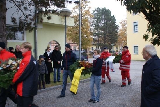 Eine Gruppe von Menschen, die rote und schwarze Jacken tragen, einige mit Blumen, gehen vor einem Gebäude mit gelber Wand. Zwei Personen in roten Uniformen tragen eine Girlande.