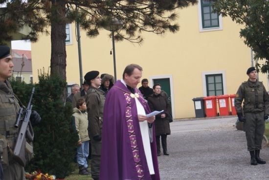 Ein Priester in einem lila Umhang steht vor einer Gruppe von Menschen, einige in Militäruniformen, in der Nähe eines gelben Gebäudes mit grünen Fenstern.