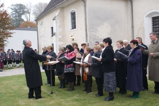 Ein Chor älterer Frauen und Männer steht auf einem Rasen vor einer Kirche und singt, während sie Notenblätter halten. Ein älterer Mann leitet sie und steht vor einem Notenständer.