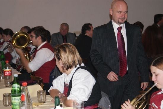 Eine Gruppe von Menschen sitzt an einem Tisch, einige spielen Musikinstrumente. Ein Mann im Anzug steht und lächelt.