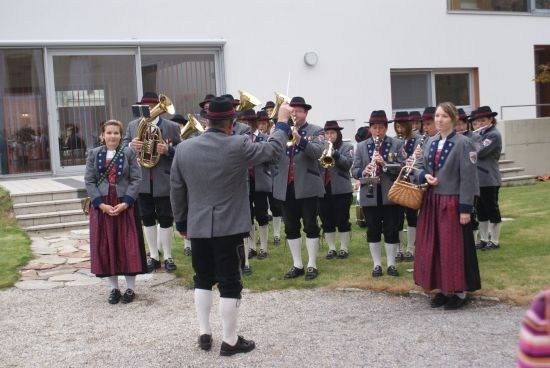 Ein Blasorchester in traditioneller Kleidung steht vor einem Gebäude, mit einer Frau im Vordergrund, die eine Tasche hält.