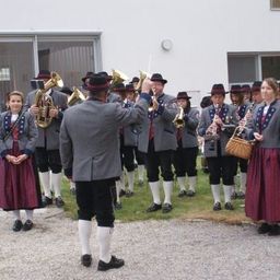 Ein Blasorchester in traditioneller Kleidung steht vor einem Gebäude, mit einer Frau im Vordergrund, die eine Tasche hält.