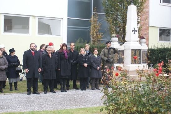 Eine Gruppe von Menschen in Wintermänteln steht vor einem Gedenkmonument mit einem Kreuz und roten Blumen.