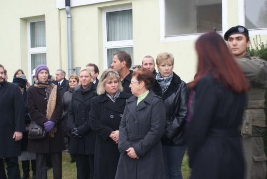 Eine Gruppe von Menschen in Wintermänteln steht vor einem Gebäude. Einige schauen in die Kamera, während andere den Boden betrachten.