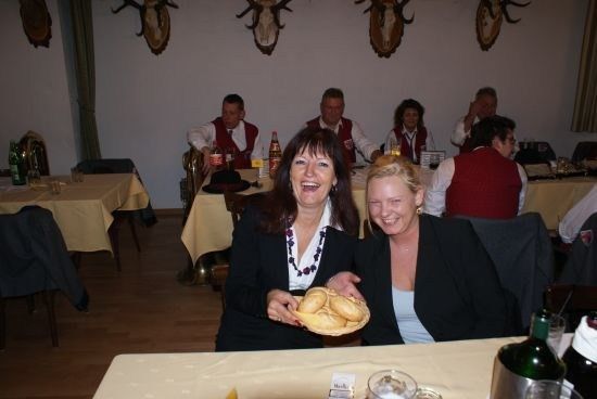 Zwei Frauen halten einen Teller mit Brot, sitzen an einem Tisch in einem Restaurant mit anderen Leuten. Der Tisch hat eine Flasche, ein Glas und einen Becher. Hinter ihnen sind andere Tische mit sitzenden Leuten und eine Wand mit Hirschköpfen.
