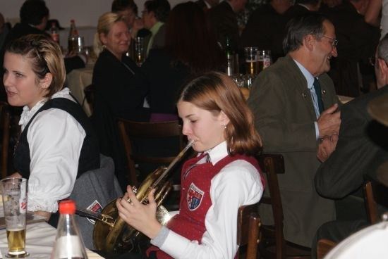 Ein junges Mädchen in einem roten Westchen spielt Trompete bei einer Zusammenkunft, umgeben von sitzenden Erwachsenen, einige lächeln und andere konzentrieren sich auf ihre Getränke.