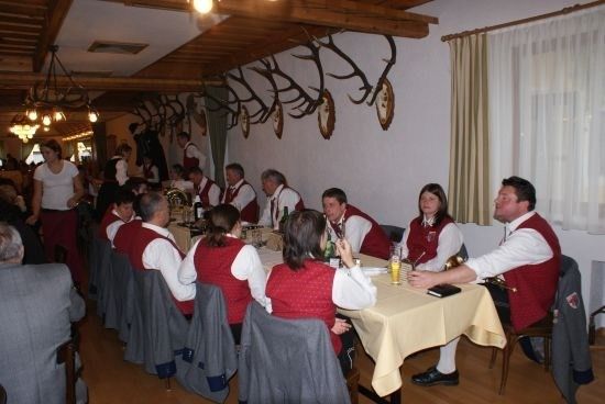 Eine Gruppe von Menschen in roten Westen sitzt um einen Tisch in einem Speisesaal. Der Raum hat Holzböden und eine Holzdecke mit Geweihen und Lampen.