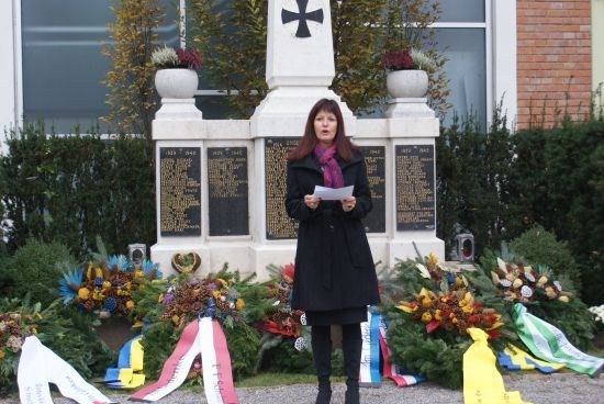 Eine Frau in einem schwarzen Mantel und Schal steht vor einem Denkmal mit Kränzen und Blumen und hält ein Stück Papier.