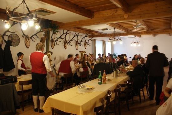 Ein Speisesaal mit Holzbalken und Geweihen an der Wand. Menschen sitzen an langen Tischen, und einige spielen Instrumente. Ein Mann in einem roten Westchen steht in der Nähe einer Trommel.