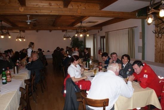 Eine Gruppe von Menschen sitzt an Tischen in einem Speisesaal, einige tragen Jacken und andere sind in formeller Kleidung. Flaschen und Gläser stehen auf den Tischen. Der Raum hat Holzbalken und einen Deckenventilator.