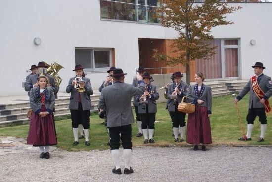 Eine Gruppe von Musikern in traditioneller Kleidung, einschließlich Hüten und langen Röcken, spielt vor einem modernen Gebäude. Sie halten verschiedene Instrumente wie Trompeten, Flöten und Tuben.