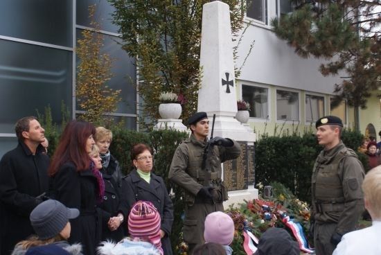 Eine Gruppe von Menschen, einschließlich Militärpersonal, steht um ein Denkmal mit Blumen und Kränzen vor einem Gebäude.