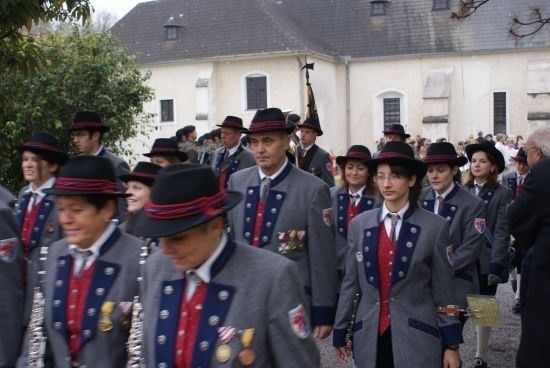 Eine Gruppe von Personen in Uniform, geschmückt mit Hüten und Medaillen, marschiert in einer Parade vor einem Gebäude.