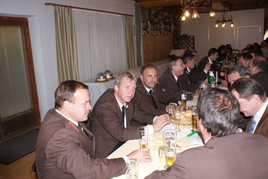 Eine Gruppe von Männern in braunen Anzügen sitzt an einem langen Tisch, trinkt Bier und führt Gespräche. Der Raum hat Holzwände und eine weiße Decke.