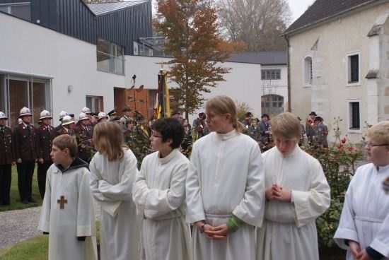 Eine Gruppe von Kindern in weißen Gewändern steht vor einem Gebäude, wahrscheinlich einer Kirche, mit einigen Erwachsenen in Uniform hinter ihnen. Sie nehmen wahrscheinlich an einer religiösen Zeremonie teil.