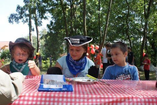 Drei Kinder in Kostümen sitzen an einem Picknicktisch mit Essen und Besteck. Eines trägt einen Piratenhut, ein anderes ein Halstuch und das dritte eine Brille. Bäume und Menschen sind im Hintergrund.