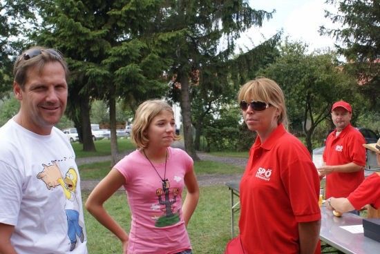 Eine Gruppe von Menschen in roten und weißen T-Shirts steht draußen. Eine Frau in einem rosa T-Shirt schaut etwas an. Ein Mann in einem weißen T-Shirt lächelt. Dahinter sind Bäume und ein Tisch.